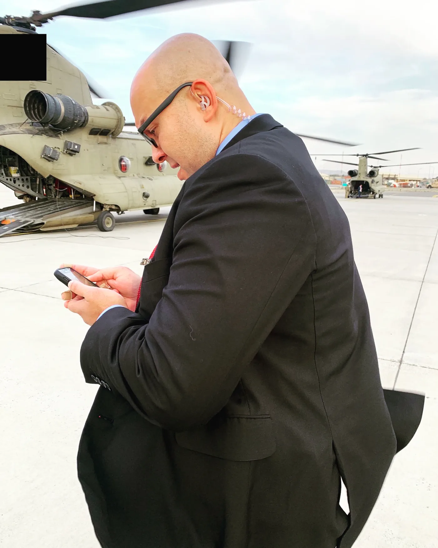 A protective agent in a suit with a clear-coil earpiece checks a phone on a flight line beside military helicopters.