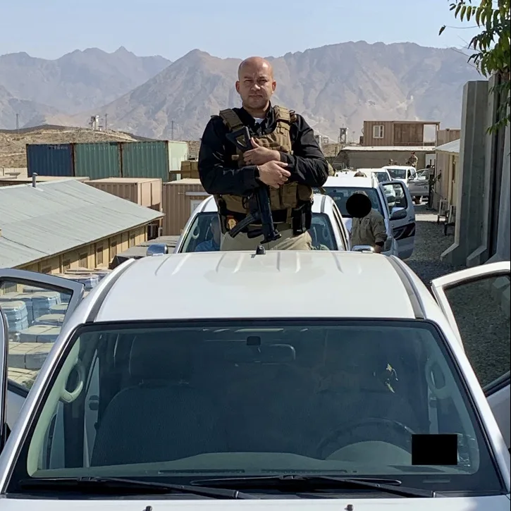 A security detail standing with vehicles on a compound road in mountainous terrain.